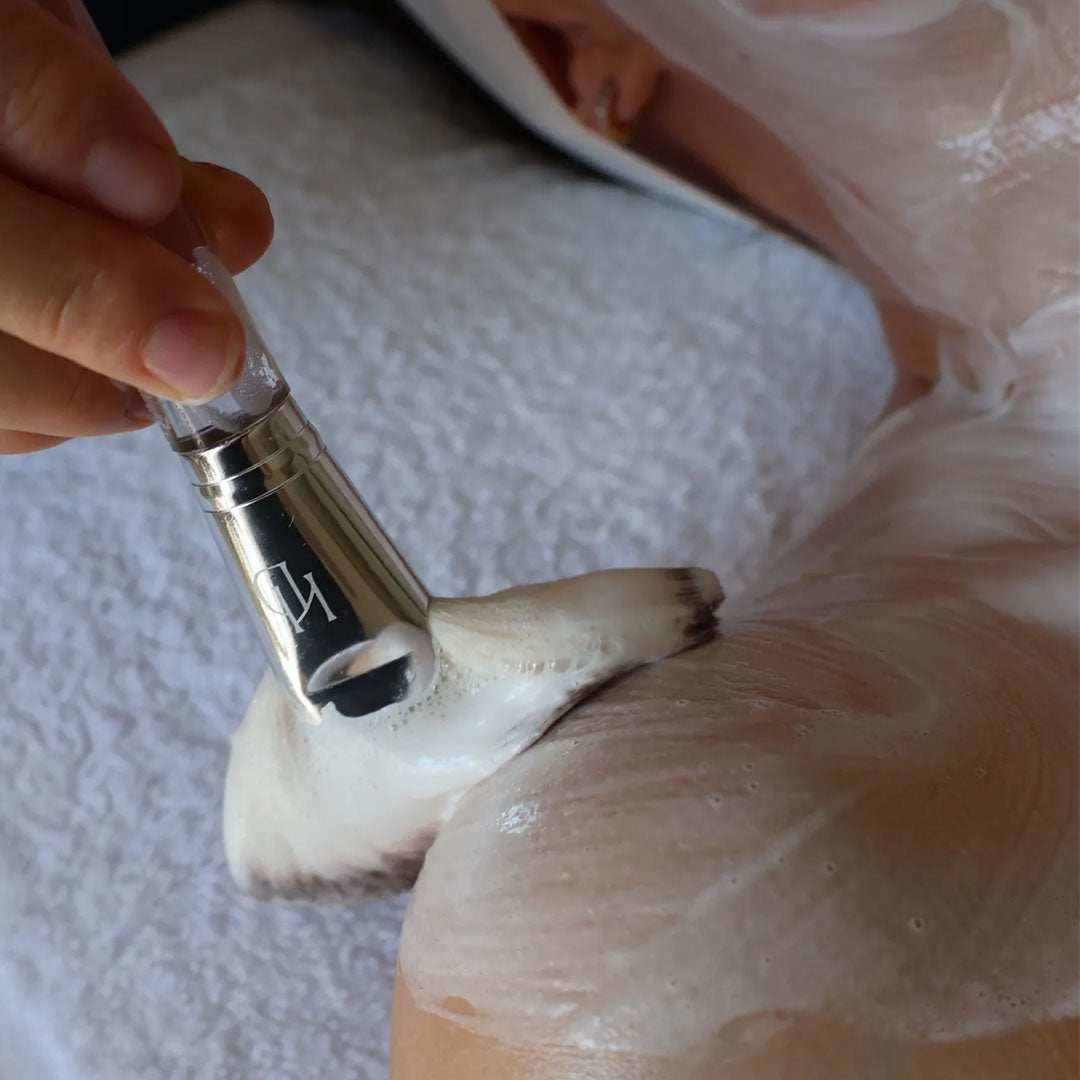 The Fluff Brush with clear handle and ultra-soft white bristles applying creamy white facial product on skin during skincare routine