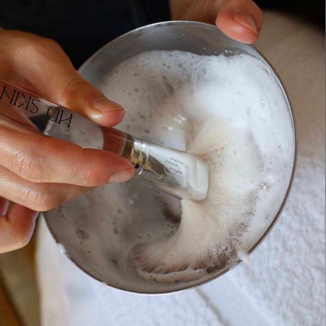 The Fluff Brush with a clear durable handle and ultra-soft white bristles creating a foamy lather in a silver bowl.